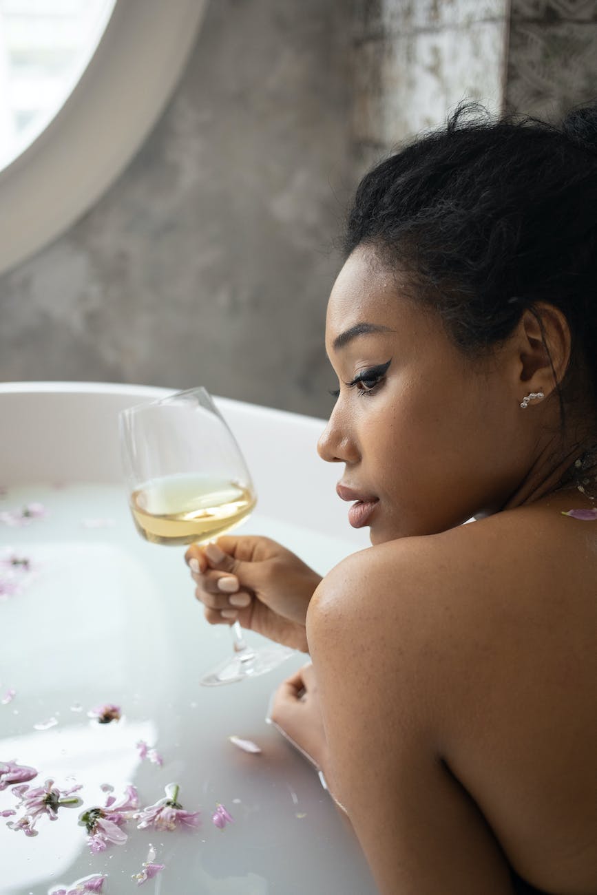 Imagen de una bella mujer sosteniendo una copa de vino blanco, dentro de una bañera, llena de pétalos de flor, en un ambiente relajante.